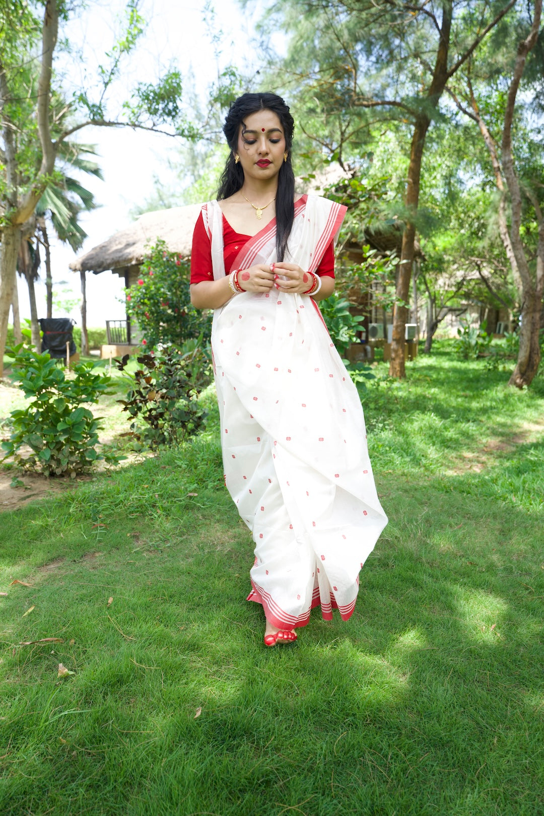 Woman in a white saree with red blouse standing on grass with trees in the background