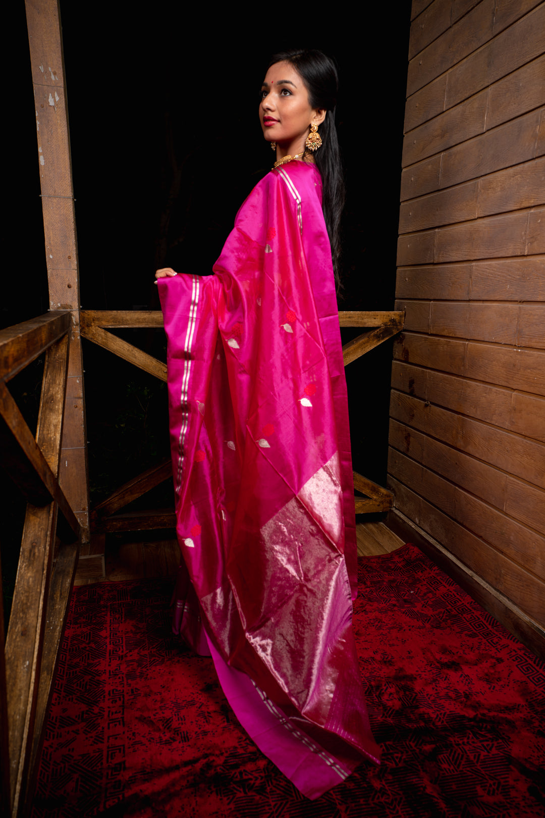 Woman in a bright pink saree with white patterns standing against a wooden wall.