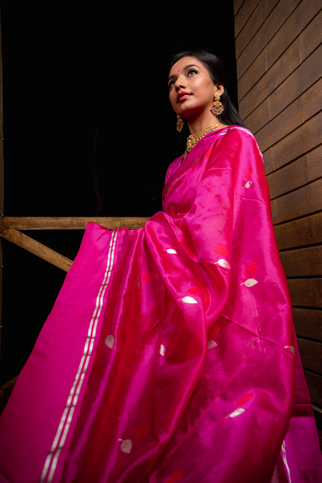 Woman wearing a bright pink saree with white patterns against a dark background