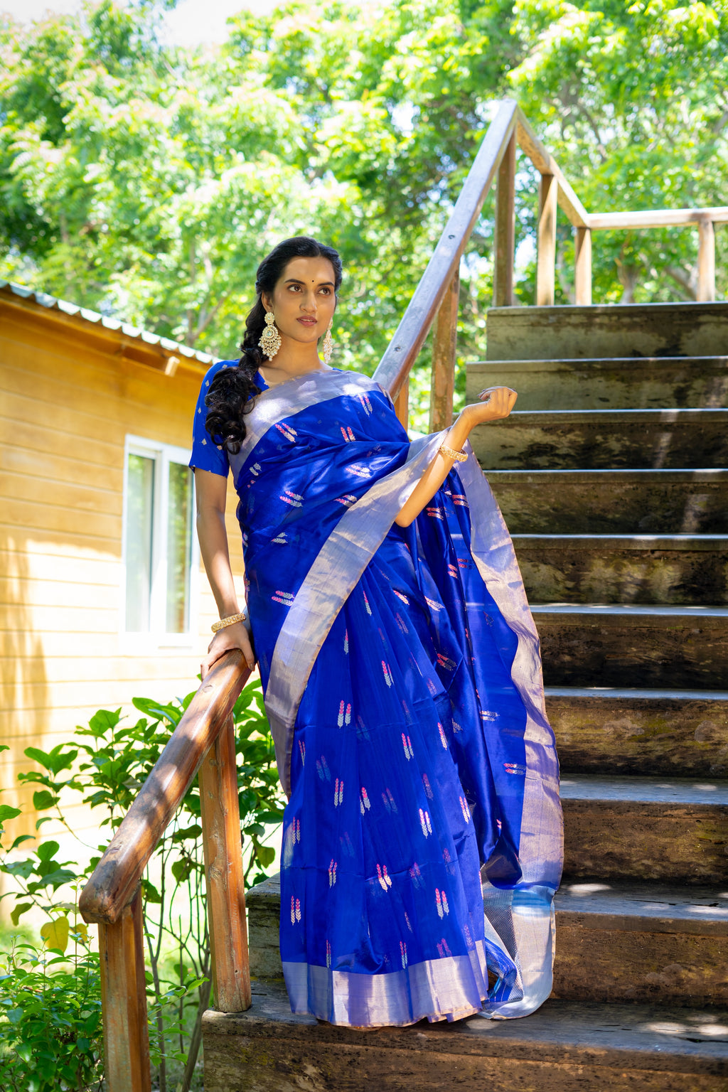 Woman in a blue saree standing on wooden stairs with greenery in the background